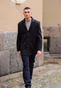 Mand med kort hår går på brostenbelagt gade iført sort frakke, grå sweater og marineblå bukser nær en stenmur.