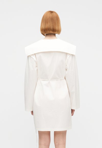 Άτομο με κοντά κόκκινα μαλλιά που φοράει λευκό φόρεμα με μακριά μανίκια και φαρδύ γιακά, εμφανιζόμενο από πίσω με απλό φόντο.