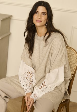 Hellbeige gestrickter Pullover mit gewelltem Ausschnitt und zarten Perlenakzenten. Weiße Spitzeärmel weisen filigrane Muster auf. Lockere Passform, kombiniert mit beigen Hosen.