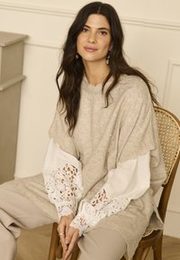 Lys beige strikket sweater med bølgemønstret halsudskæring og delikate perleaccenter. Hvide blondeærmer med intrikate mønstre. Afslappet pasform, parret med beige bukser.