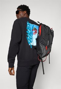 Man in een zwarte sweatshirt met blauwe grafiek op de achterkant, zwarte broek, donkere zonnebril en een zwart-rode rugzak over één schouder.