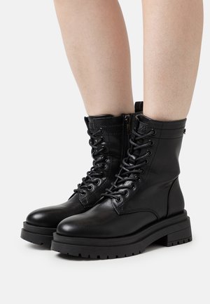 Schwarze, hochgeschnürte Lederkampfstiefel mit Schnürsenkeln und seitlichen Reißverschlüssen, getragen an nackten Beinen vor einem schlichten weißen Hintergrund.