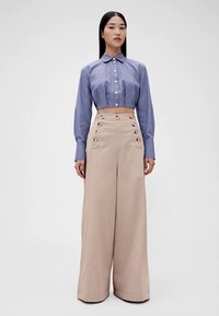 Camicia blu a righe tagliata, con dettagli di bottoni, abbinata a pantaloni beige a vita alta con gamba larga e bottoni decorativi sui lati.
