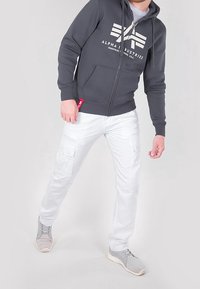 Uomo che indossa una felpa con zip Alpha Industries grigio scuro, pantaloni cargo bianchi e sneakers grigie, in piedi contro uno sfondo chiaro e semplice.