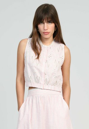 Femme aux longs cheveux bruns portant un haut sans manches rayé rose et blanc avec des boutons et une jupe assortie, mains dans les poches, fond neutre.