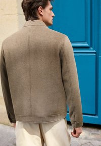 Braune Wolle-Mischjacke mit strukturiertem Kragen, glatter Textur und Nahtdetails, kombiniert mit hellen Hosen vor einem blauen Hintergrund.