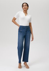 Hoge taille blauwe denim jeans met zijknopen, wijde pijpen ontwerp en lichte vervaging. Gecombineerd met een aansluitend, geribbeld wit polo shirt.