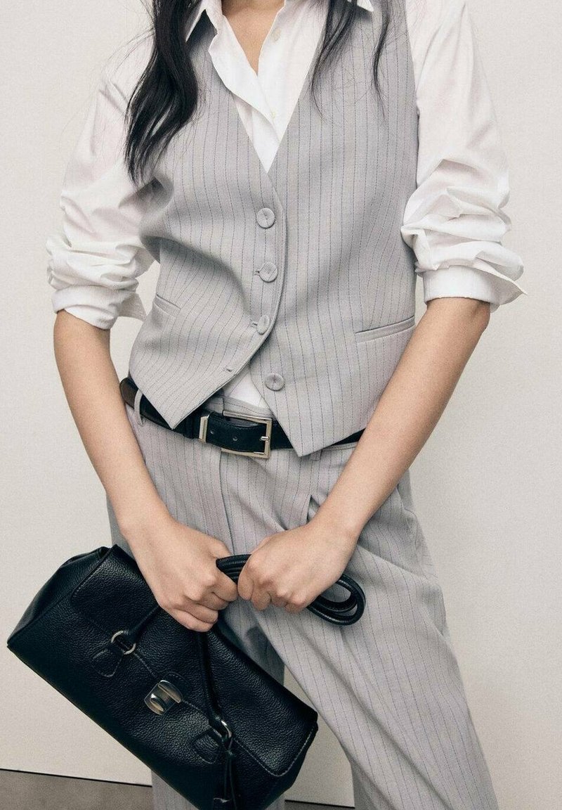 Femme portant un gilet et un pantalon gris à fines rayures avec une chemise blanche aux manches retroussées, tenant un sac à main en cuir noir par les anses.