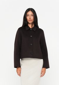 Brun ullkort jakke med knappelukking, vide ermer og krage. Glatt tekstur og minimalistisk design.