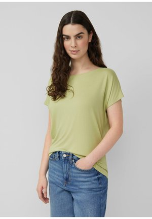 Frau mit langen, welligen braunen Haaren, die ein hellgrünes Kurzarmshirt und blaue Jeans trägt, mit einer Hand in der Tasche, stehend vor einem grauen Hintergrund.