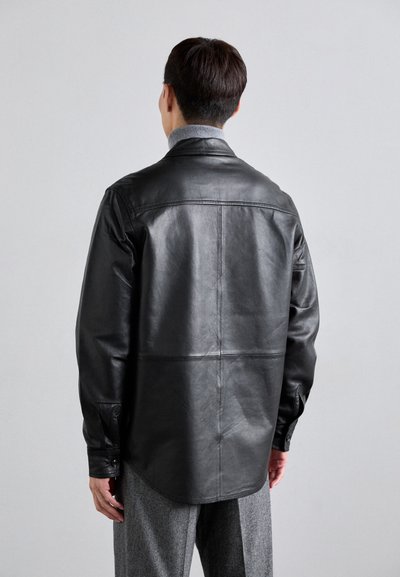 Svart läderjacka med en slät textur, böjd nederkant och knappärmar, lager på lager över en grå tröja. Bakvy.