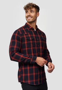 Camisa de franela en rojo y azul marino con un patrón a cuadros, mangas largas, cierre de botones en la parte delantera y dos bolsillos en el pecho. Textura suave con un corte amplio.