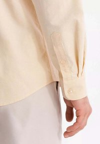 Helle gelbe Button-up-Bluse mit abgerundetem Saum, langen Ärmeln und Einknopfabschnitten. Der Baumwollstoff zeigt eine subtile Textur.