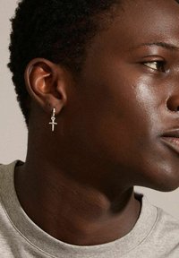 Orecchino ad anello in argento con pendente a croce. Il design presenta una finitura lucida e pende da un piccolo gancio. Indossato da un modello maschile con pelle scura.