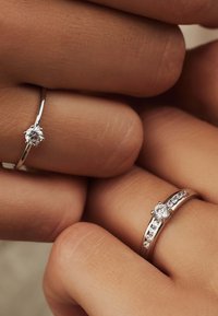 Deux bagues en argent sur des doigts : l'une présente un diamant rond central, l'autre a plusieurs petites pierres le long de la bande.