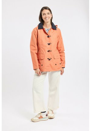 Femme souriante, portant une veste orange à boutons à bascule, un pantalon blanc et des baskets beiges, debout devant un fond blanc uni.