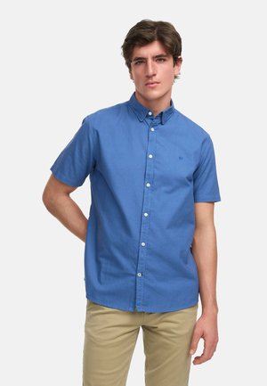 Ung mand med kort brunt hår iført en blå kortærmet skjorte med knapper og beige bukser, stående mod en ensfarvet baggrund.