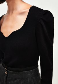 Czarna prążkowana bluzka z długim rękawem, głębokim dekoltem w serek i bufkami na ramionach, zestawiona z szarą spódnicą w paski z wysokim stanem i dwoma guzikami.