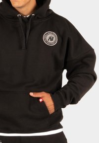 Zwarte hoodie met een rond logo patch, trekkoordkap, voorzak en een rode labelaccent op de zak. Zachte stoftextuur.