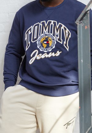 Mężczyzna noszący granatową bluzę z kapturem z logo "Tommy Jeans" oraz grafiką globu, zestawioną z beżowymi spodniami z małym podpisem Tommy.