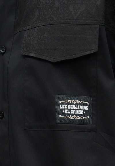 Kapsa na černé košili s texturovaným vzorovaným klopou a přišitou štítkem s nápisem "LES BENJAMINS EL GRINGO" s ozdobným ornamentem.