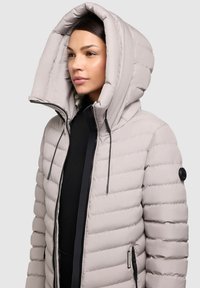 Polstret beige jakke med en stor hætte, frontlynlås, sorte detaljer og sidelommer. Vatteret struktur hele vejen igennem.