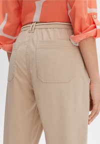 Beige Baumwollhose mit elastischem Bund, ausgestattet mit einer rechteckigen Gesäßtasche. Leichte Textur sichtbar.