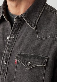 Primo piano di una persona che indossa una giacca di jeans nera sbiadita con bottoni a pressione e una tasca sul petto con un piccolo etichetta rossa Levi's.