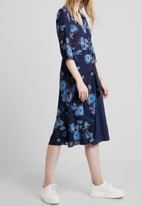 Mörkblå blommig klänning med blå blommor och prickar, med en utsvängd fåll och ärmar som går till tre fjärdedelar, i kombination med vita sneakers.