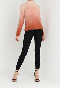 Femme portant un pull tout doux ombré passant du rose clair à la rouille, un pantalon skinny noir et des sandales à talons noirs.