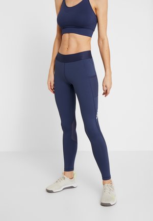 Kobieta ubrana w granatowy biustonosz sportowy i legginsy z bocznymi kieszeniami, zestawione z jasnymi butami sportowymi, stojąca na białym tle.
