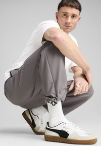 Joggers gris ajustados con puños elásticos, acompañados de zapatillas blancas con acentos negros y suelas marrones. Calcetines blancos visibles.