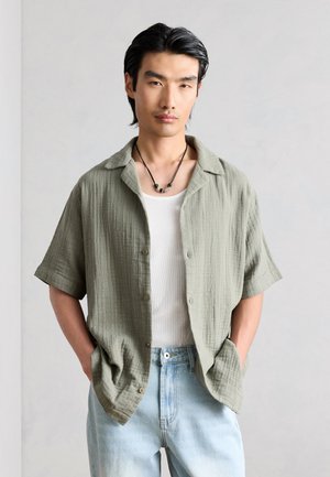 Man draagt lichtgroen kort mouw knoophemd over een wit tanktop, lichtblauwe spijkerbroek en zwarte ketting met kralen, handen in de zakken.