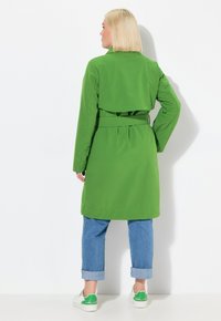 Impermeabile verde a maniche lunghe con vita cinturata, pieghe posteriori e colletto ampio, abbinato a jeans blu con risvolto e scarpe con dettagli verdi.