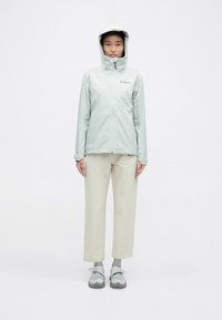 Ljusgrön vattentät jacka med huva, som har en frontdragkedja och logotyp, kombinerad med ljusbeige byxor och grå skor.