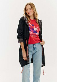 Sieviete, ģērbusies melnā capucīnā ar raibu oderi, sarkanu grafisku kaķa t-kreklu un gaiši ziliem džinsiem, stāv pret baltu fonu.