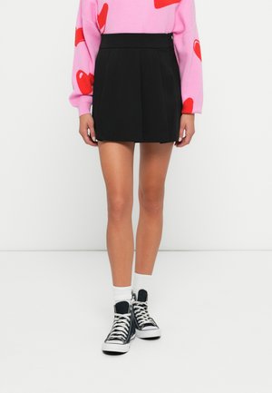 Sort plisseret mini-nederdel med en glat tekstur, båret med sorte høj-top sneakers og hvide sokker. Baggrunden er helt hvid.