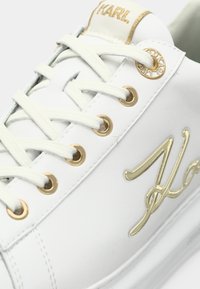 Pantofii sport albi prezintă piele netedă, glezne aurii și detalii cu șireturi. Logo-ul auriu accentuează lateralul și limba, având un design cu talpă plată.