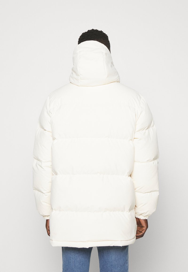 Doudoune blanche avec capuche, présentant un design matelassé et des poignets élastiques. Le tissu paraît lisse avec un léger brillant.