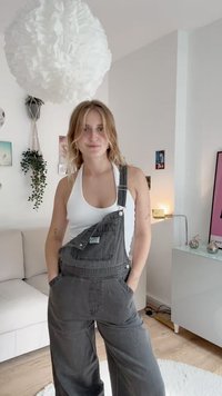 Gray denim overalls with wide legs, front pocket, adjustable straps, and a white tank top underneath. Bright indoor setting.