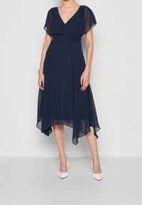 Marinblå chiffongklänning med V-ringning, korta fladdrande ärmar och asymmetrisk nederkant, matchad med vita högklackade skor.