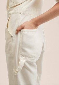 Světle krémový overal s přední kapsou a bočními kapsami, vyrobený z denimu s viditelným švem a volným střihem.