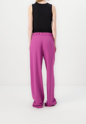 Persoon die een mouwloos zwart topje en wijde paarse broek met hoge taille draagt, staande met het gezicht naar een witte muur gericht.