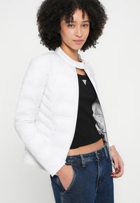 Weiße Steppjacke mit rundem Kragen, gesteppter Textur und Reißverschluss vorne, kombiniert mit einem schwarzen Tanktop und hoch taillierten blauen Jeans.