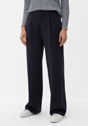 Person iført sorte vidde bukser, hvide sneakers og en sort-hvid stribet langærmet top, stående mod en ensfarvet hvid baggrund.