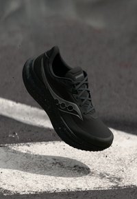 Zwarte Saucony hardloopschoen met een gegraveerde zool, zwevend boven asfalt gemarkeerd met witte lijnen.