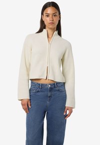 Sweater blanc court à fermeture éclair avec texture côtelée et col montant, associé à un jean en denim bleu taille haute. Design simple, quincaillerie minimale.