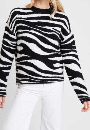 Femme portant un pull duveteux à manches longues à rayures zèbre noires et blanches, avec un pantalon blanc, debout devant un fond clair uni.