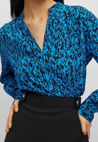 Kvinna som bär en blå och svart mönstrad blus med lång ärm inknäppt i en svart skjørt med hög midja och knapppdetaljer.