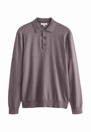 Homme portant un polo mauve à manches longues, un jean bleu et des chaussures marron à lacets, debout avec une main dans la poche.
